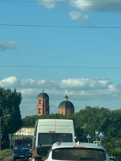 The image depicts a street scene in Заброды, Russia, on a sunny day with a clear blue sky dotted with white clouds. In the background, two church bell towers with dark domes and golden crosses rise above a dense line of green trees. The left tower is taller and made of red brick with arched openings.

In the foreground, a traffic jam is visible. Several vehicles are present, including a white van directly behind the viewer's perspective, a white car to the right of the van, and a blue KAMAZ truck further down the road. The truck has a phone number and "KAMAZ" printed on its side. Road signs are visible on the right side of the road, indicating speed bumps. A white wall runs along the left side of the road, partially obscured by trees. The overall scene suggests a typical day with traffic and religious landmarks in a Russian town.
