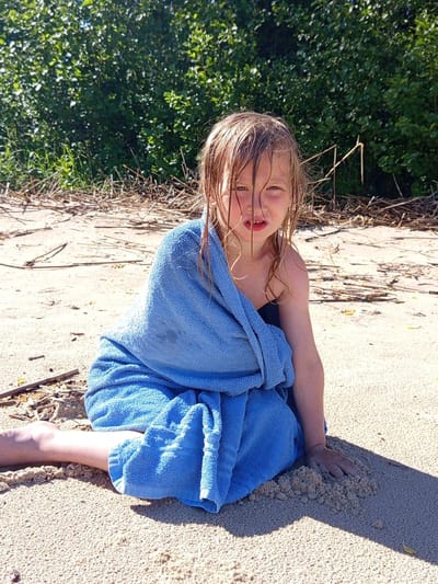 The image depicts a young, fair-skinned child, approximately 5-8 years old, seated on a sandy shore in Podborov'e, Russia, on a bright, sunny day. The child, likely female, has wet, light-colored hair (blonde or light brown) matted and partially obscuring her forehead and cheeks. Her expression is somewhat neutral, with her mouth slightly open. She is wrapped in a large, light-blue terrycloth towel, which covers her torso and legs; a dark swimsuit strap is visible on her right shoulder beneath the towel. Her right hand is partially buried in a small mound of sand directly to her right, while her left leg extends forward, partially covered by the towel. The immediate environment consists of light-brown sand with scattered dry twigs and natural debris. In the background, a dense line of vibrant green foliage, appearing as thick bushes or trees, defines the edge of the sandy area. Distinct shadows are cast on the sand by the child and towel, indicating direct sunlight from the upper right.