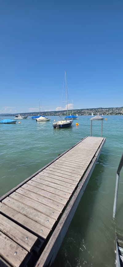 A wooden pier extends into a tranquil, greenish body of water under a clear blue sky. Several boats are moored in the water, some with blue covers and one a white sailboat with its mast standing tall. In the distance, a shoreline is visible with buildings and trees, suggesting a town or city. The pier has a metallic railing on the right side. The scene appears to be in Oberrieden, Switzerland, given the context provided. The water is calm with gentle ripples. The overall atmosphere is peaceful and summery.