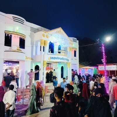 This evening image captures a bustling public event at the "চরফ্যাসন শিল্প ও বানিজ্যমেলা।" (Charfasson Industry & Commerce Fair) in Charfasson, Bangladesh. A prominent, two-story white building with ornate gold embellishments and classical-style columns serves as the central feature, illuminated by a mix of colorful green, blue, and pink lights.

A large crowd of people, including men and women, fills the foreground and surrounds the building. Many women are seen wearing traditional attire like hijabs or burqas, while men are dressed in casual wear. Attendees are actively moving, congregating, and entering the building, suggesting a lively and well-attended event.

To the right of the main building, a "টিকেট কাউন্ট" (Ticket Counter) is visible, indicating an "প্রবেশ মূল্য: ২০/-" (Entry Fee: 20/-). Further right, a larger sign provides more details: "চরফ্যাশন মাসব্যাপী ও বাণিজ্য মেলা/প্রদর্শনী-২০২৬ইং" (Charfasson Month-long Industry & Commerce Fair/Exhibition - 2026). It also mentions an "অস্থায়ী পুশ কন্ট্রোল রুম" (Temporary Push Control Room) and lists the organizers as "আয়োজক: জেলা প্রশাসক, ভোলা" (Organizer: District Commissioner, Bhola) and "বাস মালিক সমিতি, ভোলা।" (Bus Owners' Association, Bhola), confirming the location context and the nature of the event as a month-long trade fair.

The scene is brightly lit by streetlights, some wrapped in pink/purple decorative lights, and string lights draped across the fairgrounds, contrasting with the dark night sky. The ground near the entrance shows some scattered papers and debris, typical of a busy public gathering. The overall atmosphere is vibrant and festive, characteristic of a local fair or exhibition.