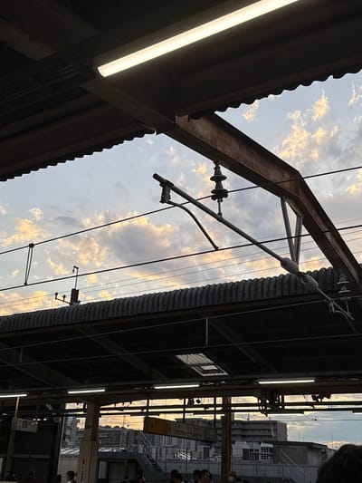 This is a medium shot, eye-level photograph of a train station platform in Funabashi, Japan, as viewed from beneath a covered structure. The sky, filled with scattered, fluffy clouds, is visible through the gaps in the station's roof. The clouds are illuminated by what appears to be late afternoon or early evening light, casting a warm glow on their edges.

Dominating the foreground are the dark, corrugated metal roof of the platform shelter and the supporting wooden and metal beams. Overhead, electrical wires for the train system are strung across the scene, held by insulators attached to metal poles and structures. One prominent metal beam, likely part of the station's infrastructure, angles diagonally across the upper right portion of the image.

Below the main roofline, another corrugated metal roof is visible, also casting shadows. In the distance, beyond the train station structures, buildings of varying heights and architectural styles can be seen, suggesting an urban environment. Some signs with Japanese text are visible on poles, indicating platform numbers or directions.

In the lower portion of the image, a group of people are present on the platform, some standing and some walking. They are mostly obscured by shadows and the station's structure, but their presence indicates an active transportation hub. The overall impression is one of a functional urban setting at the end of the day, with the natural beauty of the sky providing a backdrop to the man-made structures.
