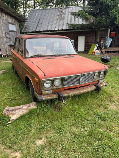 An aged, red Lada sedan sits outdoors in a grassy area, partially obscured by tall grass and foliage. The car appears to be in a state of disrepair with rust and missing parts visible. The front of the car is facing toward the top-right corner of the image. Behind the car, a weathered wooden building with a metal roof is visible. Miscellaneous items are stacked near the building, including a car tire, boxes, and what appears to be a grill. The location is outdoors, overgrown, and gives the impression of being in a rural or semi-rural area. The angle of the photograph suggests it was taken at eye-level, possibly from a slightly elevated position. There is no indication of it being Philippeville, Belgium.