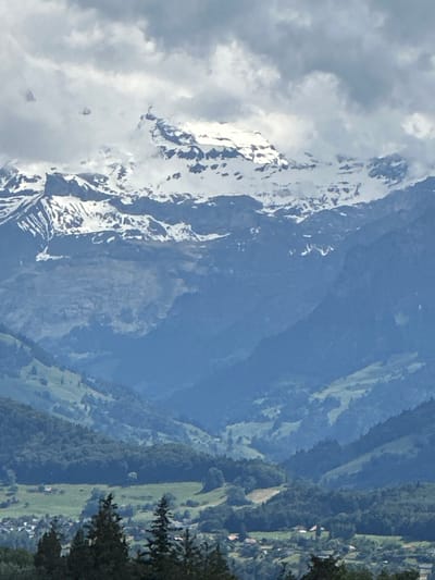 The image depicts a mountainous landscape with snow-capped peaks in the background, under a cloudy sky. In the foreground, rolling hills covered in green vegetation and trees descend into a valley. A small village with red-roofed houses is visible nestled within the valley. The scene suggests a rural or semi-rural environment, likely in the Swiss Alps. Given the context provided about the city of Thun, Switzerland, this landscape is likely a view from or of the mountainous regions surrounding Thun.