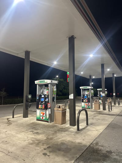 The image depicts a 7-Eleven gas station, likely in Homosassa Springs, Florida, United States. There are multiple gas pumps visible under a canopy, each displaying 7-Eleven branding. The pumps feature digital screens and various buttons. Trash receptacles are positioned near the pumps. Bike racks are present in front of each of the gas pumps. The ground is a concrete pavement. The scene is illuminated by the canopy lighting, and it appears to be nighttime or early morning, as the sky is dark. There are trees and greenery behind the gas pumps.