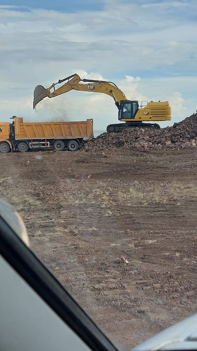 An excavator is dumping rocks into a dump truck on a construction site. The scene is in the city of Saran, Kazakhstan. The sky is cloudy with patches of blue. The ground is dirt and rocks. There is a white vehicle partially in the foreground.