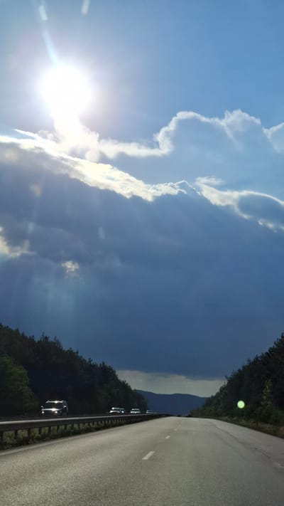 An asphalt highway in Tserovo, Bulgaria, is viewed from the perspective of a vehicle traveling along it. The highway is flanked by dense green pine forests on either side. In the distance, rolling hills are visible. The sky above is a dramatic mix of bright sunlight breaking through dark, heavy clouds. Several cars are visible on the highway, appearing as distant headlights or silhouetted forms. The road is marked with white lines. The overall impression is of a dynamic and atmospheric landscape, possibly during or just after a storm.