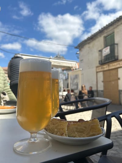 The image depicts an outdoor dining scene in a public square or street in El Espinar, Spain, under a clear blue sky with scattered white clouds. In the foreground, two glasses filled with golden-colored beer, each topped with a layer of white foam, rest on a white outdoor table. To the right of the beer glasses, a small white plate holds two triangular slices of Spanish tortilla. A dark metal outdoor chair frame is partially visible in the lower right.
In the midground, several blurred figures are seated at other outdoor tables, with one individual partially visible on the far left wearing a light green quilted jacket. The background features light-colored, possibly whitewashed, buildings with tiled roofs. On the right, a two-story building displays a green-shuttered window and a dark metal balcony with a "SE VENDE" (For Sale) sign. Its ground floor has closed wooden doors. In the center-background, an architectural structure with a dome or cupola is visible, along with an awning or sign reading "El Casino." Overhead, utility lines traverse the frame against the sky. The scene is brightly lit by natural sunlight.