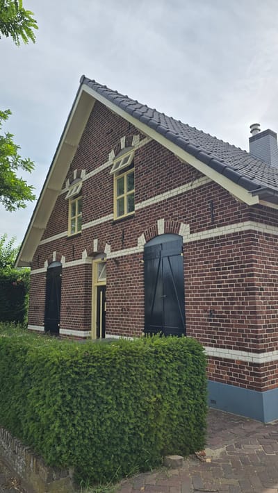 A brick house with a dark tiled roof is pictured in the city of Zeddam, Netherlands. The house has a light-colored trim around the roofline and white horizontal accents in the brickwork. There are two windows on the upper level with yellow frames and shutters, which are open. On the lower level, there are two large dark wooden doors with shutters, and a smaller entrance door with a yellow frame. A neatly trimmed green hedge runs along the front of the house, and a brick pathway is visible in the foreground. A chimney with a grey cap is visible on the roof. The sky is overcast.