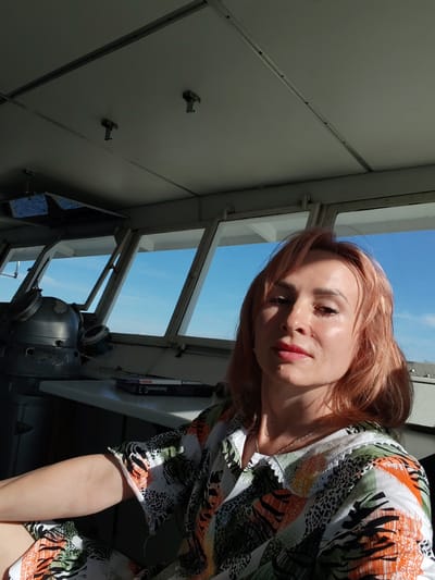A woman with light pink hair and makeup is taking a selfie in what appears to be the bridge of a ship, with windows showing a bright blue sky. She is wearing a colorful shirt with a pattern of black, white, orange, and green. Sunlight is streaming in from the right, illuminating her face and shoulder. To her left, a large, vintage-looking ship's compass is visible. Several books are stacked on a surface in front of the windows. The ceiling of the bridge is visible in the upper part of the image, with what look like hooks or fixtures attached. The setting suggests it is daytime, likely sunny. The woman's expression is neutral, but the lighting creates a warm and somewhat reflective mood. There is some text visible on one of the books, "Е ОБУЧЕНИЕ". The location is identified as Dargun, Russia.