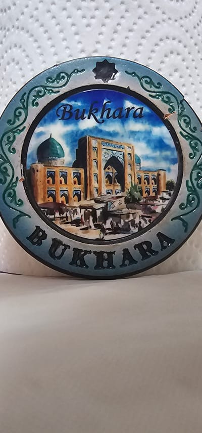 The image shows a circular decorative plate featuring a scene of a historic city and the name "BUKHARA" inscribed on the outer rim. The central image depicts architectural structures with domes and intricate patterns, characteristic of Central Asian or Middle Eastern cities. Small figures of people are visible in the foreground, suggesting a bustling marketplace or public area. The plate has a decorative border with green, ornate designs. The background of the plate is a painted sky with clouds. The plate itself is set against a textured white surface, likely a paper towel. It is not possible to determine the specific location within Karshi City, Uzbekistan, from the image provided, as the plate depicts Bukhara.