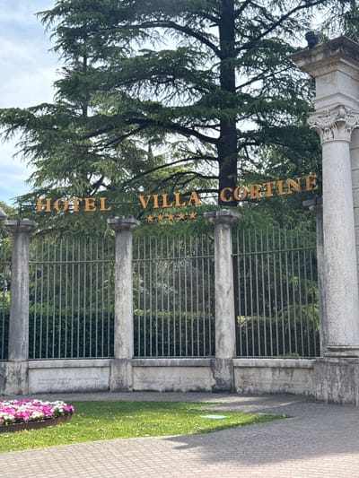 The image shows the entrance to Hotel Villa Cortine in Sirmione, Italy. A large, ornate sign with gold lettering spells out "HOTEL VILLA CORTINE" above a metal fence. The fence is supported by four stone pillars. Behind the fence, lush green trees and foliage are visible, and a large evergreen tree dominates the background. The sky above is cloudy. In the foreground, a bed of colorful flowers sits on a patch of green grass next to a paved walkway. The stone base of the fence has engraved text, but it is not legible. No people are visible in the image.
