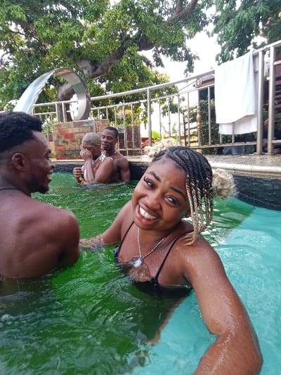 Four people are in a swimming pool with murky green water. A woman in a black swimsuit with braided hair smiles directly at the camera. A man with short curly hair is to her left, facing away from the camera. Two individuals stand near a stainless steel fountain feature in the background; one is a blonde woman covering her face, and the other is a man shirtless. A white towel is hanging off a metal fence near the edge of the pool. Lush trees and foliage are overhead.