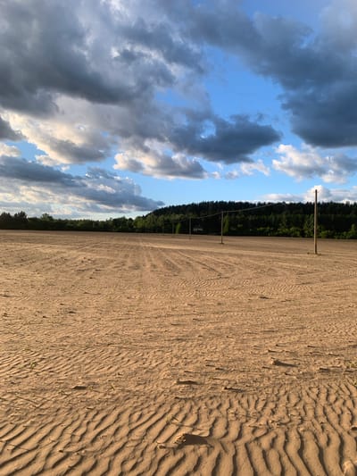 The image shows a field in Varviškė, Lithuania. The ground is sandy with wavy patterns. Parallel lines run across the field, suggesting tire tracks. A line of trees and wooden poles are visible along the horizon. The sky is partly cloudy with a mix of blue and gray clouds. There are no people visible in the scene.