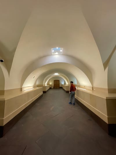 A person stands in the middle of a dimly lit, arched corridor in Strelna, Russia. The corridor features a vaulted ceiling with recessed lighting and a skylight, as well as a cream-colored wainscoting along the lower walls. The floor is made of dark stone tiles. The person, wearing a red jacket, grey pants, and blue slippers, is facing away from the camera, looking down the corridor towards a closed wooden door at the far end. A security camera is visible on the left wall near the entrance of the corridor.