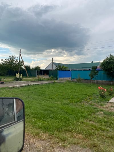 The image depicts a residential area in Krylovskaya, Russia, on a cloudy day. The foreground shows a patch of green grass and a side-view mirror of a vehicle, reflecting a person wearing a mask. Beyond the grass, a paved path runs horizontally, bordered by concrete curbs. To the left of the path is a lawnmower and a tall plant with white flowers. Further back, a green metal fence encloses a property.

Behind the fence, a long, low-slung building with a green metal roof is visible, possibly a residential house or a farm structure, with an open-sided extension. Another green fence, a portion of which is bright blue, runs along the front of this building. Trees and shrubs are scattered throughout the scene, with power lines strung between utility poles. In the distance, the sky is dominated by heavy, gray clouds, with patches of lighter sky visible. To the right, a bush with several pink and orange roses is in bloom, and a portion of a concrete structure is visible.