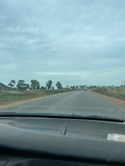 View from inside a vehicle traveling on a paved road lined with trees in Kujama, Nigeria. The sky is overcast. A few vehicles are visible in the distance ahead. The vehicle's interior and windshield are partially visible.