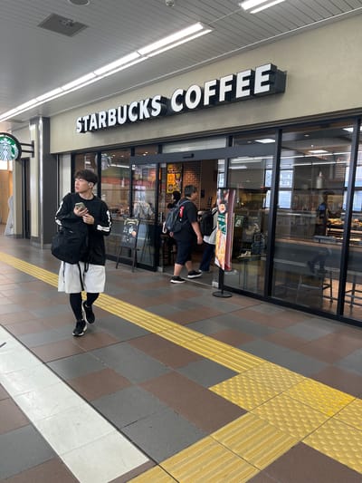 The image depicts a lively indoor commercial corridor, likely within a train station or shopping complex in Shinjuku, Japan. A Starbucks Coffee establishment dominates the center of the frame, identifiable by its prominent black and white "STARBUCKS COFFEE" signage and a green circular siren logo affixed to the building and reflected in the glass doors.

In the foreground, a young East Asian male walks from left to right across a multi-toned tiled floor composed of brown and grey squares. He is dressed in a black long-sleeved shirt with white stripe details on the sleeves, light grey shorts worn over black leggings, and black athletic shoes. He carries a black backpack on his left shoulder, holds a light-colored smartphone in his right hand, and wears white wireless earbuds. His gaze is directed upwards and to the right. A yellow tactile paving strip, featuring both parallel raised lines and circular raised dots, runs horizontally across the floor in front of the Starbucks, indicating a path for visually impaired individuals. A wide white line is painted on the floor in the lower left.

The Starbucks entrance consists of glass doors allowing visibility into the well-lit interior, which features a counter area, display cases with food items, and multiple menu boards, some displaying Japanese text. Several individuals are visible inside the store or entering/exiting. A tall, slender promotional stand with an image of food and Japanese text, along with the English words "GOOD START MORNING," stands directly in front of the Starbucks entrance. A smaller black A-frame chalkboard sign is positioned to the left of the entrance. The ceiling is paneled with recessed rectangular fluorescent lights and includes an access panel and a small dome-shaped camera or sensor.