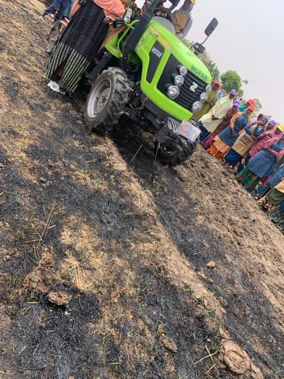 A green WoFan tractor with a large rear tire is parked on a dirt field in Ringimawa, Nigeria. The field has been recently burned and is covered in black ash and charred grass. A group of people are standing behind the tractor and appear to be watching or waiting. The person in the foreground is wearing a black and white dress and is standing behind the tractor on the left side. The tractor's front tire is visible in the background, along with the people. The ground is uneven, and there are traces of mud. The tractor is facing forward, but its tires are slightly turned to the right. The weather is cloudy and there is a small amount of vegetation in the background.