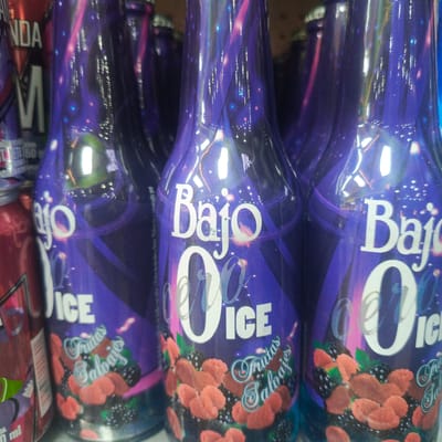 This is a close-up, eye-level shot of several bottles of "Bajo 0 ICE" fruit-flavored beverage, a product of Empresas Polar, lined up on a shelf in what appears to be a retail environment. The bottles are purple with a psychedelic design featuring swirling lines, stars, and depictions of raspberries and blackberries. The text "Bajo 0 ICE" is prominently displayed in white lettering, with "Frutas Salvajes" (Wild Fruits) written below it. The bottles are reflective, showing highlights from overhead lighting. The background is blurred, but suggests a commercial setting, possibly a supermarket or convenience store in Los Teques, Venezuela. The image focuses on the product packaging, highlighting its vibrant colors and branding. No people are visible in the frame.