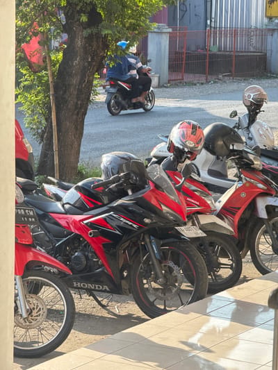 In Kedawung, Indonesia, several motorcycles are parked along a paved area with a tiled walkway. The motorcycles are predominantly red and black, with one lighter colored scooter visible. Helmets are placed on some of the motorcycles, including a red and black one with "KYT" branding and a beige helmet. In the background, a person on a motorcycle wearing a blue jacket and helmet rides away from the viewer. A tree with green foliage stands to the left, and a fence with red bars encloses an area behind the parked vehicles. The scene is illuminated by sunlight, casting shadows on the ground.
