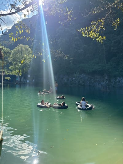 People float on inner tubes in a clear, turquoise-tinted lake. The bright sunlight casts long reflections across the water, creating a shimmering effect. Lush greenery borders the lake, with trees and dense foliage rising up the hillside. A few wooden structures are visible along the shoreline. The perspective is slightly tilted, adding to the sense of immersion in the scene.