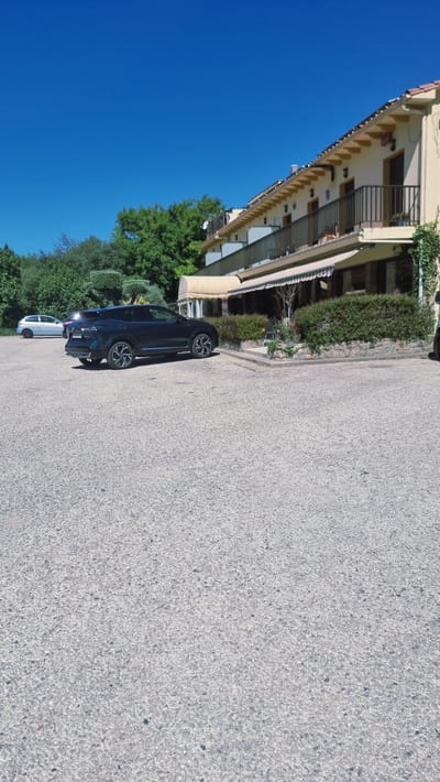 The image depicts a parking lot in front of a building that appears to be a restaurant or hotel. The building has a light-colored facade with multiple balconies and awnings. The parking lot is surfaced with gravel, and a dark-colored SUV is parked prominently in the foreground. Another smaller, lighter-colored car is visible further back in the lot. Lush green trees and vegetation surround the building, and the sky is clear and blue. The scene appears to be set on a sunny day. There are no individuals clearly visible in the image.