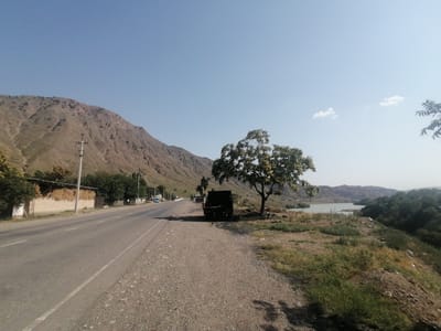 The image captures a roadside scene near Tash-Komur City, Kyrgyzstan, on a clear day. A paved road runs diagonally from the lower left to the middle of the frame, bordered by a gravel shoulder. To the left of the road, a low brick building with a covered roof is filled with hay bales. A utility pole stands in front of it. Further along the road, several trees line the left side.
On the right side of the road, a large, bare tree stands prominently. To its right, a dark, boxy vehicle, possibly a truck or utility vehicle, is parked on the gravel. Beyond the tree and vehicle, a body of water, likely a reservoir or lake, is visible, with hills and mountains in the background. The landscape is rugged and arid, with sparse vegetation on the slopes. The sky is a clear blue with a few wispy clouds. There are no people visible in the image.