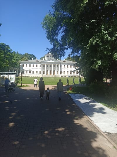 The image depicts the manor house of Ostafievo, a museum-estate in Russia, under a clear blue sky. The grand white neoclassical building features a portico with large columns and a domed rotunda. In the foreground, a paved pathway leads towards the house, lined with trees casting dappled shadows. Several people are walking on the pathway, including a person pushing a stroller, a couple walking hand-in-hand with a child, and other individuals walking in various directions. A large white tarp is laid out on the grass beside the pathway. The environment is a park-like setting with a well-maintained lawn in front of the manor.