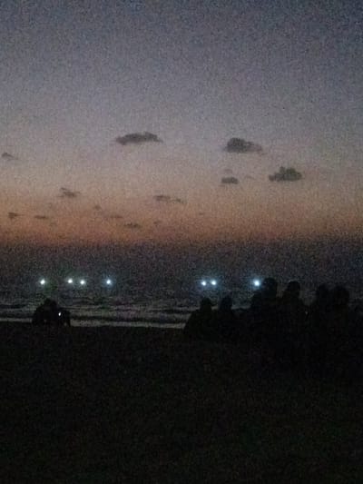 The image displays a twilight or nighttime scene on a beach in Al-Mawasi, Palestinian Territories. The sky transitions from a faint orange-pink hue near the horizon to a darker blue-grey overhead, populated by several small, dark clouds. The dark ocean surface is visible, with subtle wave patterns near the shore. Six distinct, bright white lights are present on the water, appearing as three pairs horizontally across the mid-ground. The foreground consists of a dark, indistinct beach area. Silhouetted figures are visible on the beach: a smaller, possibly crouching or seated figure or group is located on the left near the water, while a larger group of multiple figures is seated or gathered on the right. The scene is characterized by low light conditions and digital image noise.
