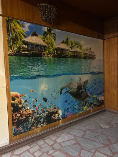This image depicts an interior space in Sushitsa, Bulgaria. A large, framed wall mural dominates the scene, presenting a split-level view of a tropical environment.

The upper portion of the mural illustrates an above-water scene featuring several thatched-roof bungalows built on stilts over calm, turquoise water. These structures are nestled amidst lush green palm trees under a bright blue sky with scattered clouds. The water surface shows subtle ripples.

The lower portion of the mural depicts an underwater scene. Vibrant coral formations, in shades of red, orange, and brown, populate the foreground and midground. Numerous tropical fish of varying species, colors, and sizes swim among the coral. Three grey dolphins are visible towards the right-center of this section: two smaller dolphins are positioned above and slightly ahead of a larger, darker dolphin.

The mural is bordered by wooden paneling along the top and right side, which extends to the ceiling. A multi-tiered crystal chandelier hangs from the wooden ceiling above the mural. The floor consists of a mosaic-style pattern made of irregularly shaped, light-colored stone or tile pieces interspersed with reddish-brown grout lines. No people are visible.