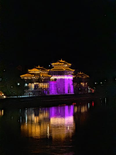 The image shows a structure illuminated in gold and purple lights at night, reflected in what appears to be water. The structure is multi-tiered with a traditional architectural style, suggesting it is located in Zhuoshui, China. The surrounding environment is dark.