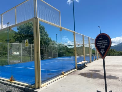 This media file depicts an outdoor padel tennis court with glass walls and a metal mesh enclosure. The court surface is a vibrant blue, marked with white lines. The surrounding area is paved with concrete, and there are patches of water on the ground, suggesting recent rain or a sprinkler system. In the background, lush green hills and trees are visible under a clear blue sky with scattered white clouds.
A prominent sign on a black pole stands to the right of the court. The sign is triangular with rounded corners and features a black and orange design. It displays text that reads: "LANDMARKER GROUP", "THE WORLD'S HIGHEST PADEL COURT AT 2,003 METERS", and "PADEL ISLAND". This suggests the location is at a high altitude.
There are no people visible in the image. The scene is set during the daytime, likely mid-morning or early afternoon, given the bright sunlight and the shadows cast. The weather appears to be pleasant and sunny, though the presence of water on the court hints at possible recent precipitation. The mountainous landscape and the "Padel Island" branding on the sign provide location cues, consistent with the provided context of Kumlistsikhe, Georgia, known for its high-altitude attractions. The overall impression is one of a modern sporting facility situated in a scenic, elevated natural environment.