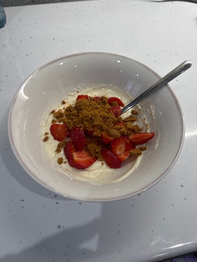 A close-up, overhead shot captures a bowl of yogurt topped with sliced strawberries and crushed biscuits. The bowl is white with a subtle pink rim, and it rests on a speckled white countertop. A silver spoon is partially submerged in the yogurt, suggesting the food is being eaten or is ready to be eaten. The lighting is bright, indicating it's likely daytime. The overall impression is one of a simple, healthy snack or dessert. There is no visible text in the image. The location context of Craigavon, United Kingdom is provided separately.