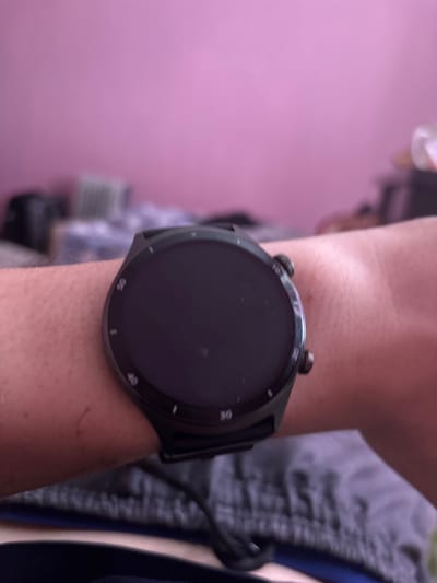 This is a close-up, waist-up shot of a person's arm wearing a smartwatch. The smartwatch is round with a black face and bezel. The bezel has markings that appear to be numbers, possibly indicating minutes or hours, with "10", "20", "30", "40", and "50" being partially visible. The watch is attached to a black strap. The person's arm is lightly tanned and shows some skin texture. They are wearing a grey garment, possibly athletic shorts, on their right side. In the background, to the left, is a blurred area of pink and white, suggesting a wall or backdrop. The lighting appears to be natural and diffused. There is no visible text on the smartwatch face. The scene is casual and domestic. The setting is likely indoors, given the soft lighting and indoor background elements. No specific time of day or weather conditions can be determined.