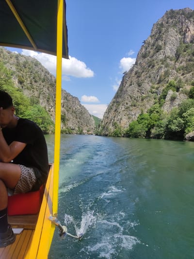 The image captures a view from a yellow boat moving through a canyon in Dolna Matka, North Macedonia. In the foreground, a person wearing a black t-shirt and patterned shorts is seated on the left side of the boat, facing away from the camera. The boat has a yellow frame and a dark green canopy. The boat is creating a wake as it travels down a green body of water. On either side of the water, steep, rocky canyon walls rise up, covered in patches of green vegetation. In the distance, more boats are visible on the water, and there appear to be structures on the far bank of the canyon. The sky above is a clear blue with scattered white clouds.