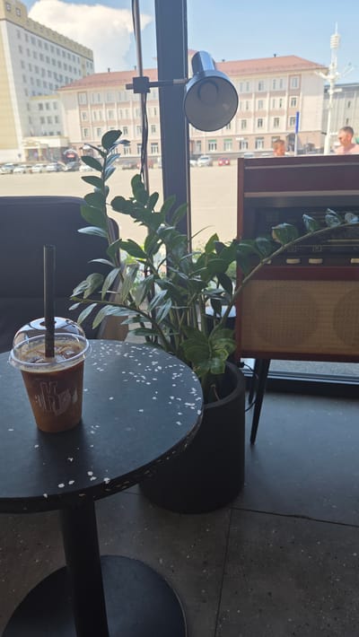 In Gorno-Altaysk, Russia, a cafe scene is depicted, focusing on a table in the foreground with a beverage and a plant. A plastic cup containing a brown iced drink with a black straw rests on a dark, circular table speckled with white. Next to the table, a potted plant with lush green leaves is visible. In the background, through a large window, a street scene unfolds with buildings and parked cars. A vintage radio with wooden accents and a speaker grille is positioned to the right of the plant. A desk lamp with a metal shade is attached to the window frame. Two men are partially visible outside, one standing and the other seated.