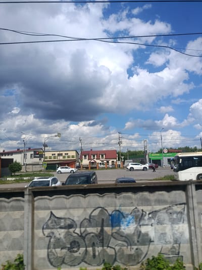 The image depicts a street scene in Elektrougli, Russia, on a partly cloudy day. In the foreground, a concrete fence is covered in graffiti. Behind the fence, a road is visible with several cars, including a white SUV, a black SUV, and two white sedans. A white bus is also present on the road. Several buildings line the street, some of which appear to be commercial establishments, with signs in Russian. The sky is a mix of blue and dark gray clouds. People are visible on the sidewalk and near the bus. The environment appears to be a typical urban or suburban setting.