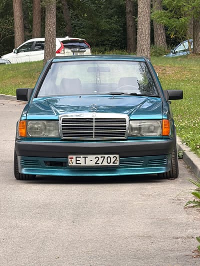 This media file shows the front of a teal-colored Mercedes-Benz 190E parked on a paved surface, likely a street or parking area, in Upeslejas, Latvia. The car is positioned diagonally to the viewer, with its front grille and headlights prominently displayed. The license plate, reading "ET-2702", is clearly visible. To the left of the car, a line of trees with rough bark stands alongside a grassy area with scattered wildflowers. In the background, another vehicle, possibly a white SUV, is partially visible, along with more trees and foliage. The lighting suggests it is daytime, and the weather appears to be pleasant. The scene is static, with no people or overt activity taking place. The overall impression is of a parked car in a semi-natural, suburban or rural setting.