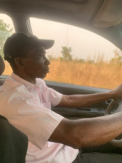 An older man drives a car. He wears a black baseball cap and a striped, short-sleeved shirt. The outside scenery consists of dry grass and a few trees visible through the car window. The man's arms are raised, holding the steering wheel. The image is shot from an angle, looking slightly up at the driver.