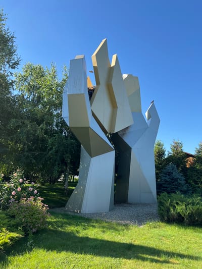 A large, abstract, metallic sculpture stands in a garden under a clear blue sky. The sculpture is composed of sharp, angular planes that are mostly silver with some beige accents. It has a dynamic, almost flame-like appearance, reaching upwards. The sculpture is situated in a well-maintained garden with lush green grass, manicured bushes, and a rose bush with pink flowers on the left side. Behind the sculpture, various trees, including birch and evergreen varieties, are visible, along with a hint of a wooden building to the far right. A small bird is perched on the top of the sculpture. The scene appears to be located outdoors, likely in a park or private garden setting. The image is taken from a medium distance, capturing the entire sculpture and its surrounding environment.