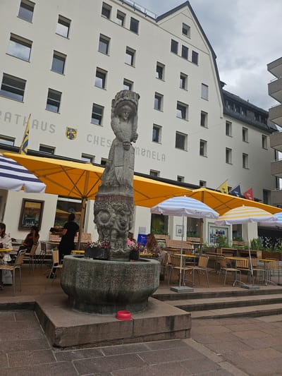 The image shows a street scene in St. Moritz, Switzerland, likely during daytime. A fountain with a stone sculpture stands in the foreground, with red lettering carved into its base. Behind the fountain, several yellow and striped umbrellas are set up, suggesting an outdoor seating area for a cafe or restaurant. Tables and chairs are arranged beneath the umbrellas, and a few people are seated, appearing to be dining or socializing. In the background, a modern building with multiple windows and some text written on its facade is visible. Flags are also displayed, one of which has the text "St. Moritz" on it. The sky is overcast, indicating cloudy weather. The overall atmosphere seems relaxed and casual, with people enjoying the outdoor space.