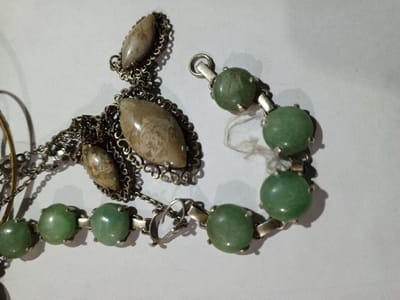 In the city of Phakeo, Laos, two distinct pieces of jewelry are displayed overlapping on a flat, light-colored surface. The first item, positioned primarily on the left and top, is a dark-toned metal necklace or pendant featuring three polished, elongated oval-shaped stones with mottled beige and brown coloration. Each stone is set within an ornate, filigree-edged bezel, with the largest stone centrally placed and two smaller stones situated above it, all connected by a fine-link chain. The second item, curving across the bottom and right, is a silver-toned metal bracelet or necklace composed of multiple uniformly rounded, light green stones, each secured in a four-prong setting and linked by small rectangular connectors. A partial clasp mechanism, including a jump ring, is visible at one end of this piece. A thin, light-colored fiber is entangled near one of the green stone settings. The scene is illuminated by direct overhead lighting, casting subtle shadows.