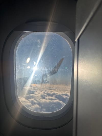 The image displays an aerial view from inside an airplane, looking out through an oval-shaped window. The window surface exhibits multiple scratches and significant glare from direct sunlight, which appears as a prominent starburst flare in the upper-left quadrant. The upward-curving tip of an airplane wing is silhouetted against the bright sky, partially obscured by the sun glare, in the upper-mid left of the external view. Below the wing, a dense and uniform layer of white cumulus clouds extends across the lower horizon. Above the clouds, the sky is a clear, light blue. The interior visible includes a gray window frame and adjacent gray paneling on the right. The scene is observed from an aircraft in flight above a cloud layer. No ground features are visible, rendering the specific location in the city of Tekirdağ, Turkey, indeterminable from this image.
