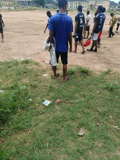 The image depicts an outdoor scene with several young men in athletic attire on a dirt field, likely a sports ground, with patchy grass in the foreground. In the background, buildings suggest an urban or semi-urban environment, possibly within Ukpor, Nigeria. The young men appear to be standing around, some in conversation, others observing. One person is seen from behind, wearing a blue and black checkered shirt. Another individual, also from behind, has a black shirt with the name "KELVIN" and the number "14" on it. To their right, another young man wears a black shirt with "FHOUX" and "7" on it. The ground is littered with various pieces of trash, including plastic wrappers and a discarded can. The lighting indicates it is daytime.
