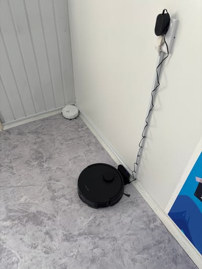 In a corner of a room with white, vertically paneled walls and a grey marble-patterned floor, a black robot vacuum cleaner is parked on its charging base. The charging base is plugged into a white electrical outlet on the wall, which also has a black adapter plugged into it. A black cord snakes down the wall from the adapter and connects to the charging base. Further back in the corner, near the wall, sits a white robot vacuum cleaner. A small blue light is visible on the black robot's charging base, indicating it is charging. To the right of the charging area, a portion of a colorful graphic poster is visible on the wall. The image appears to be taken indoors, in a domestic setting, possibly a home or apartment. The scene does not contain any information that would allow for its specific location in Vileyka, Belarus.