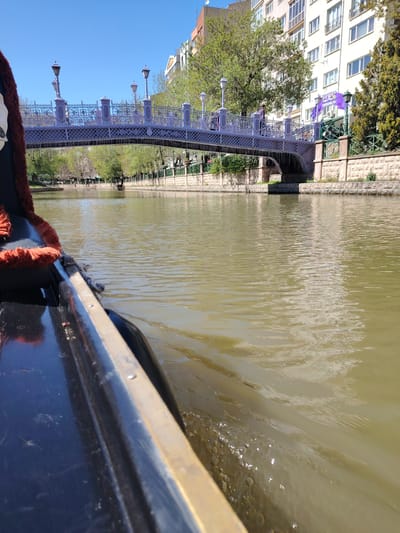 The image presents a sunlit view from a boat navigating a river or canal in Eskisehir, Turkey. The perspective is low, looking forward from the front-left side of a dark-colored boat, possibly a gondola, which features a reddish-brown fuzzy trim. Ripples and small bubbles indicate the boat's movement through the light brown-green water.
In the midground, an ornate bridge, characterized by its decorative metal railings and street lamps, spans the waterway. Several individuals are visible crossing the bridge. Beneath the bridge, another boat can be seen further along the canal.
The riverbanks are lined with stone walls and abundant greenery, including trees with fresh leaves suggesting spring or summer. On the right bank, a large, multi-story white building with numerous windows dominates the background, accompanied by other urban structures. The sky overhead is clear and a vibrant blue, indicative of a bright daytime setting.
A purple flag or banner attached to the right side of the bridge structure prominently displays the text "ESKİŞEHİR YILI" (Eskişehir Year) along with a colorful graphic, confirming the location. The scene conveys a tranquil and enjoyable experience of a canal tour.