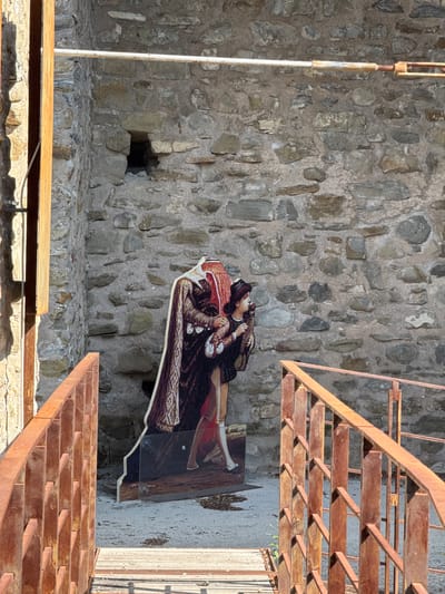 A cutout figure depicting a man in historical attire stands in front of a stone wall, partially obscured by a rusty metal railing. The figure's head is cropped out of the frame, but the elaborate cloak and the lower half of the man's body, including his legs and feet in stockings and shoes, are visible. He is shown from the waist down, with his right hand touching a bird perched on his left hand. The background is a rough stone wall with an opening to a dark space. A narrow wooden walkway with a rusty metal railing leads towards the figure. The scene is located in Dolceacqua, Italy, near the Doria Castle.