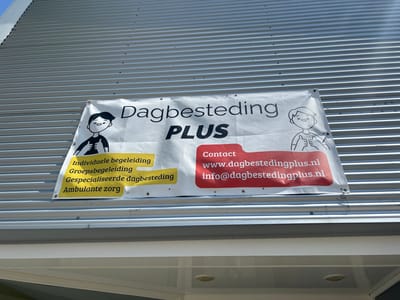 A white banner is attached to a corrugated metal wall, likely the side of a building in Lisse, Netherlands. The banner advertises "Dagbesteding PLUS" (Daycare PLUS) and lists services such as "Individuele begeleiding" (Individual guidance), "Groepsbegeleiding" (Group guidance), "Gespecialiseerde dagbesteding" (Specialized daycare), and "Ambulante zorg" (Outpatient care). Contact information for "Dagbestedingplus.nl" is also prominently displayed. Two cartoon figures, one on the left with arms raised and the other on the right giving a thumbs-up, are depicted on the banner. The overall setting appears to be an outdoor facade of a commercial or institutional building.