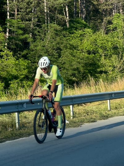 A person is riding a bicycle on a road bordered by a metal guardrail and lush green trees. The cyclist is wearing a white helmet and a light green cycling suit, with bright yellow accents on the shorts. The rider's left leg is extended, pushing down on the pedal, while the right leg is bent, completing the circular motion of cycling. Both hands are on the handlebars.
The scene appears to be set during the daytime, likely late morning or early afternoon, given the bright sunlight casting shadows on the road. The weather seems fair and warm, as indicated by the vibrant greenery and the cyclist's attire. The background consists of dense trees with some dry grass and weeds growing along the roadside. The road itself is asphalt and appears to be a paved route, possibly in a rural or semi-rural area given the natural surroundings.
The overall impression is one of athletic activity and a serene, natural environment. The focus is on the cyclist's movement and the dynamic action of riding. There is no visible text in the image. The setting, with the guardrail and trees alongside a road, suggests a cycling route in a scenic location, possibly in Bulgaria, near Prisovo, as per the location context.