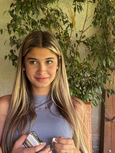 A young woman with long blonde and brown hair is seated outdoors in front of a large green plant and a textured wall, possibly in the city of Golyama Zhelyazna, Bulgaria. She is wearing a light blue sleeveless top and small pearl earrings. The woman is holding a pack of cigarettes in her left hand and appears to be interacting with a white object in her right hand, which could be a cigarette or a lighter. Her gaze is directed to her left. The background consists of dense green foliage and a light-colored, textured wall. A wooden chair back is visible behind her right shoulder.